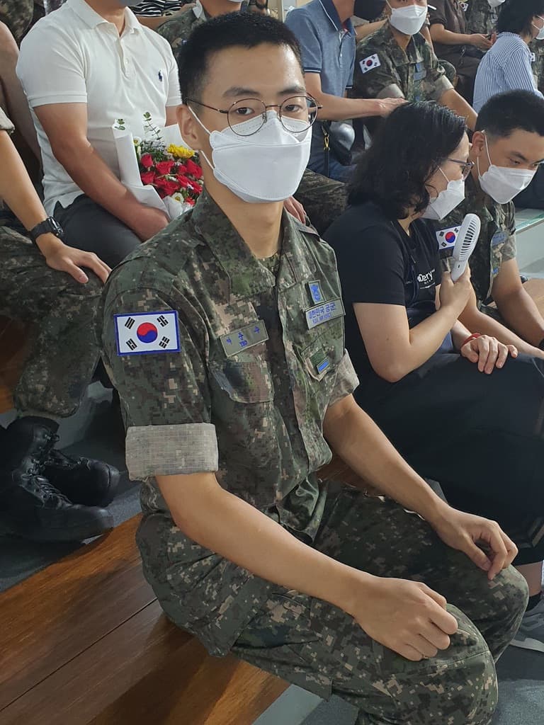 신병수료식에서 디지털 위장 군복과 마스크 차림으로 가족 옆에 앉아 있는 여준수